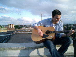 a handsome man wearing a half open blue shirt holding a acoustic guitar sitting outside on the roof of a building